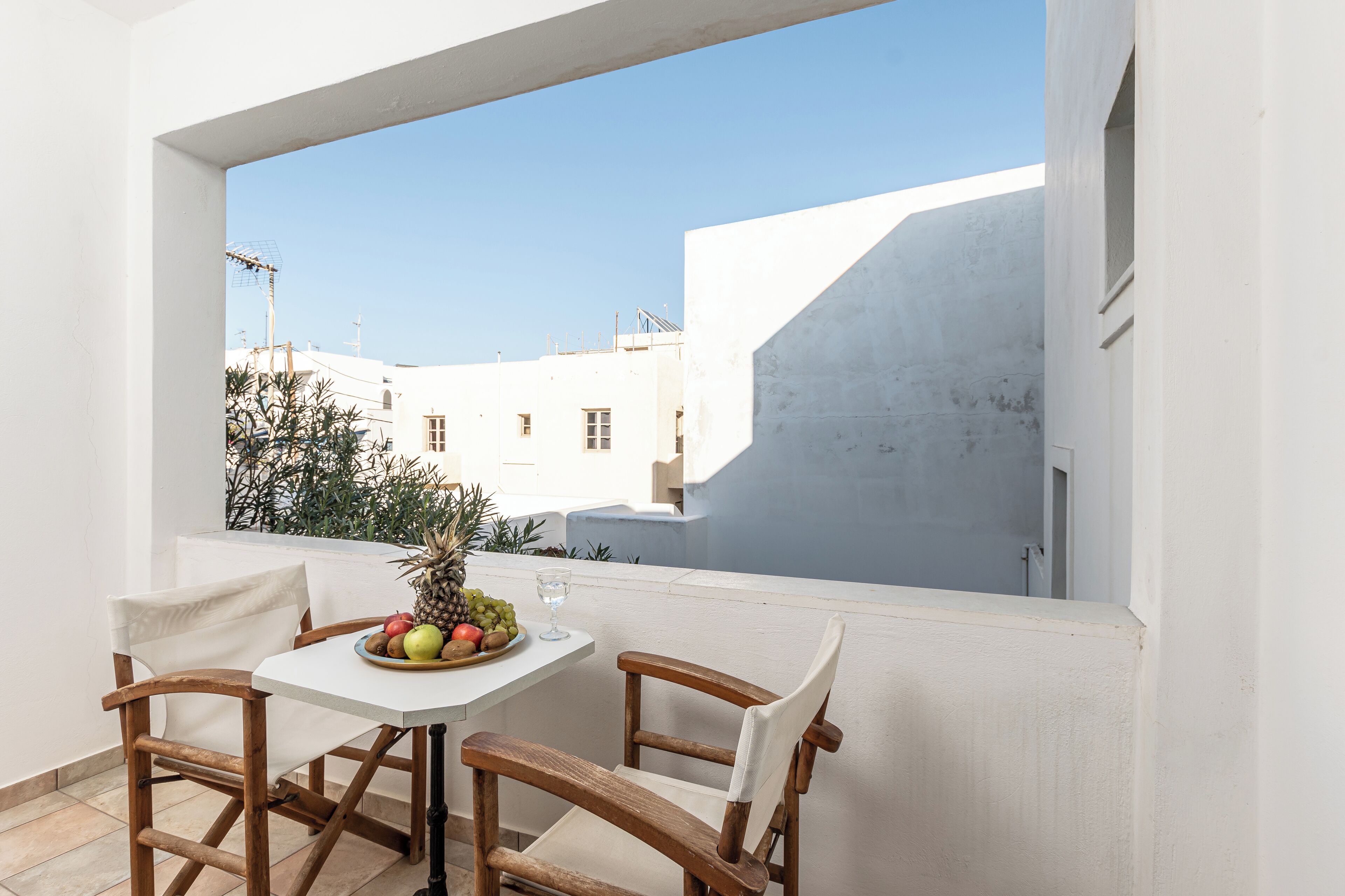 Un balcón con una mesa y sillas blancas, un frutero y una vista despejada de edificios y cielo.