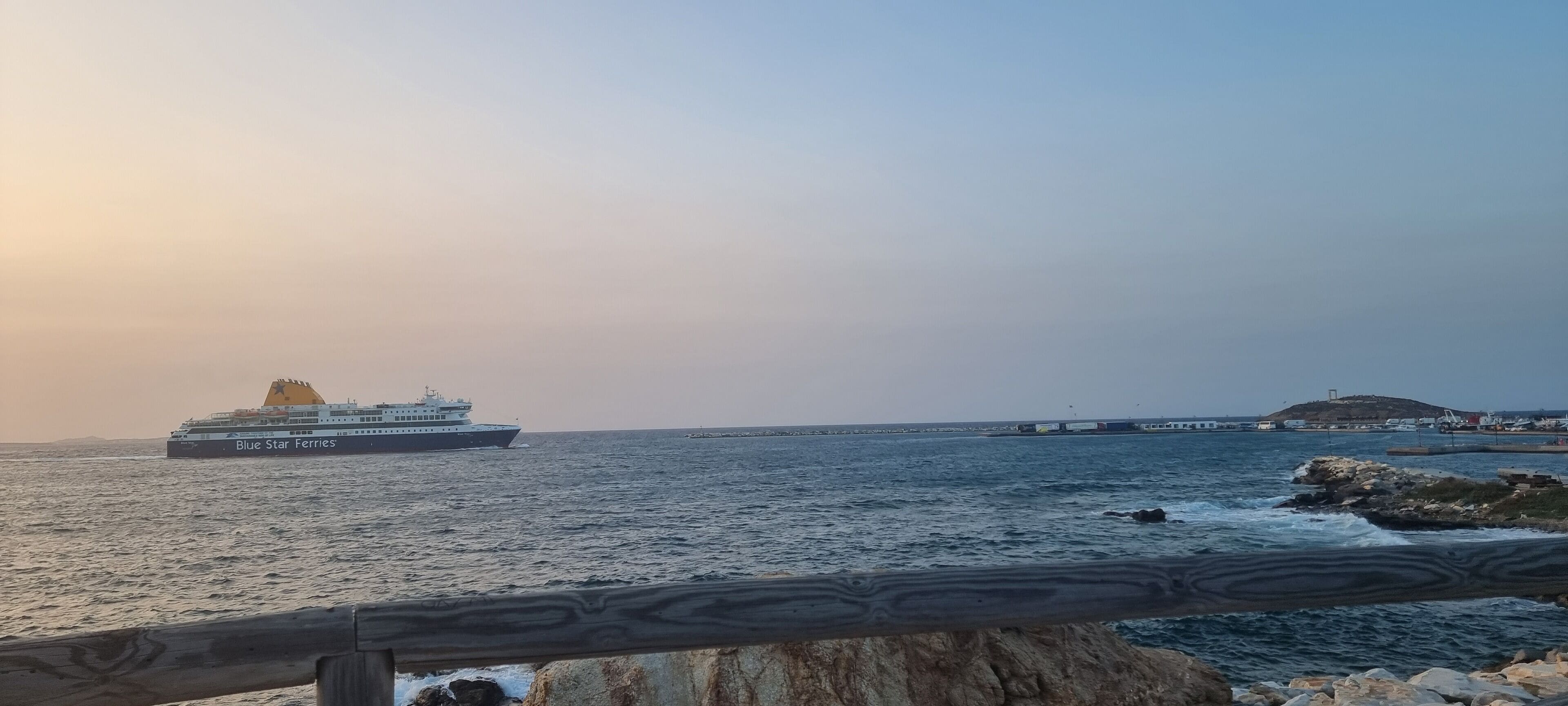Een veerboot genaamd Blue Star Ferries op het water, met een rotsachtige kustlijn en een eiland in de verte.