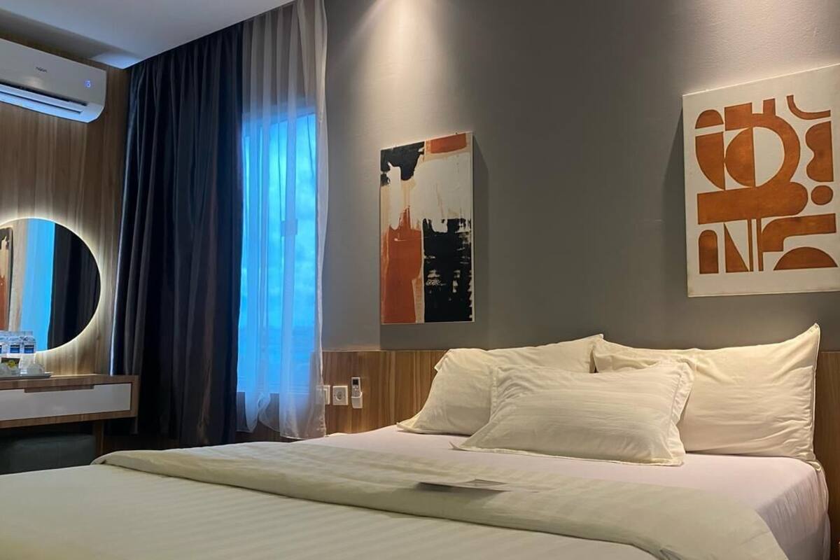 A hotel room with a bed, a bedside table with a lamp, a wall-mounted air conditioner, and a window with curtains.