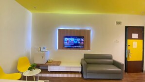 A modern living room with a flat-screen TV mounted on the wall, a grey sofa, yellow chairs, and a small white table with a plant. - Kaliban Hotel (Batam)