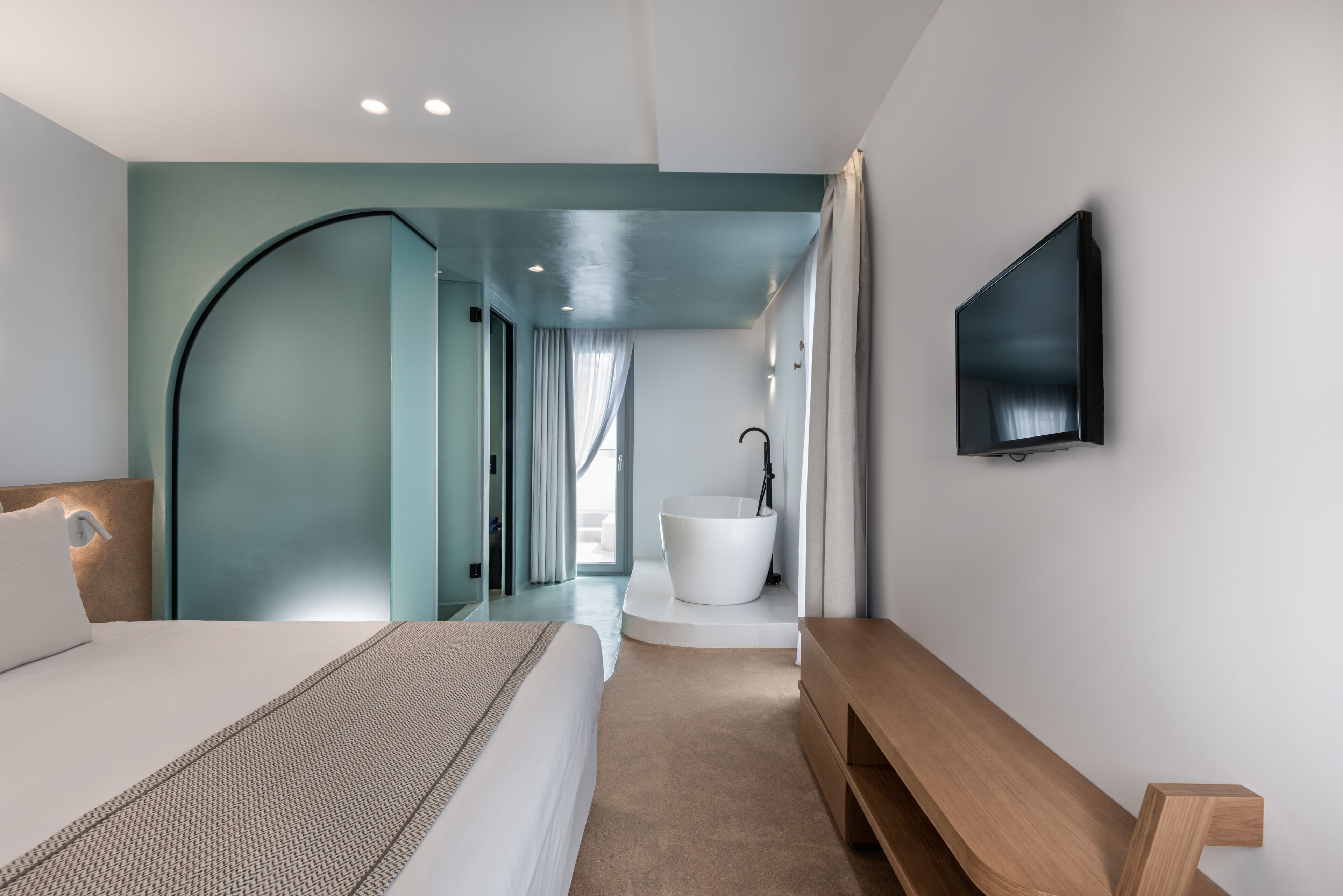 A modern hotel room with a bed, a wooden bench, a large mirror, a flat-screen TV, and a white bathtub.