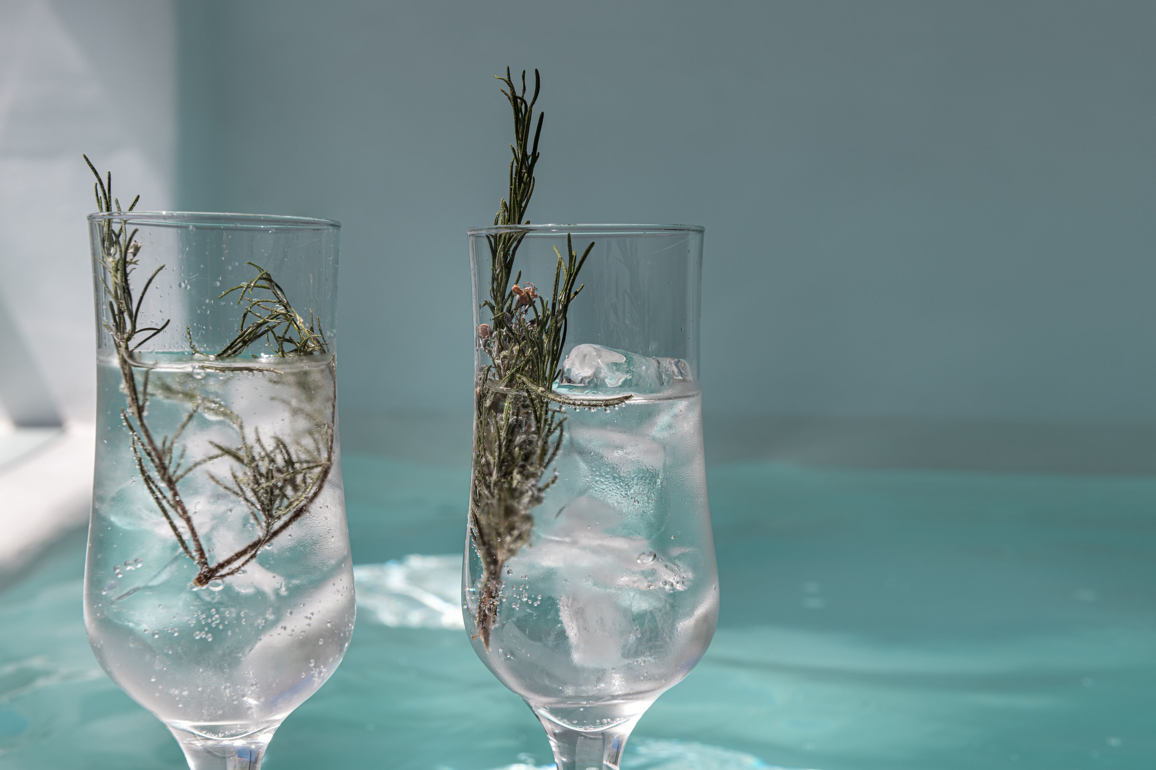Two glasses of water with rosemary branches, placed on a reflective surface.