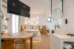 Un dormitorio moderno con cama, mesitas de noche, televisor, espejo redondo y baño con lavamanos y espejo.