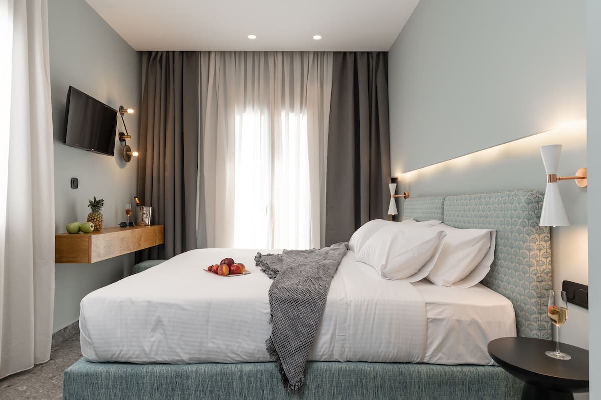 Moderni hotellihuone, jossa on suuri sänky, seinälle asennettu televisio, yöpöytä lampun kanssa ja seinälle kiinnitetty hylly, jossa on hedelmiä.