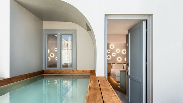 A modern indoor pool area with a wooden deck, a glass door leading to a bedroom, and circular wall lights. - Argo Boutique Hotel (Naxos)