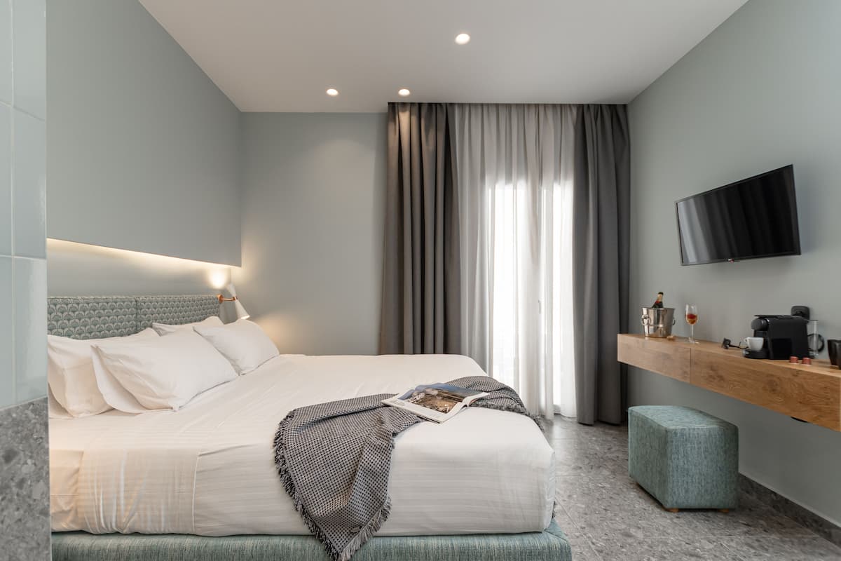 Moderni hotellihuone, jossa on siististi pedattu sänky, seinälle asennettu televisio, puinen konsolipöytä, jossa on kahvinkeitin, sekä suuri ikkuna, jossa on verhot.