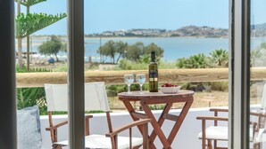 A wooden table with a bottle of wine, two glasses, and a bowl of fruit on a balcony overlooking a beach and sea. - Argo Boutique Hotel (Naxos)