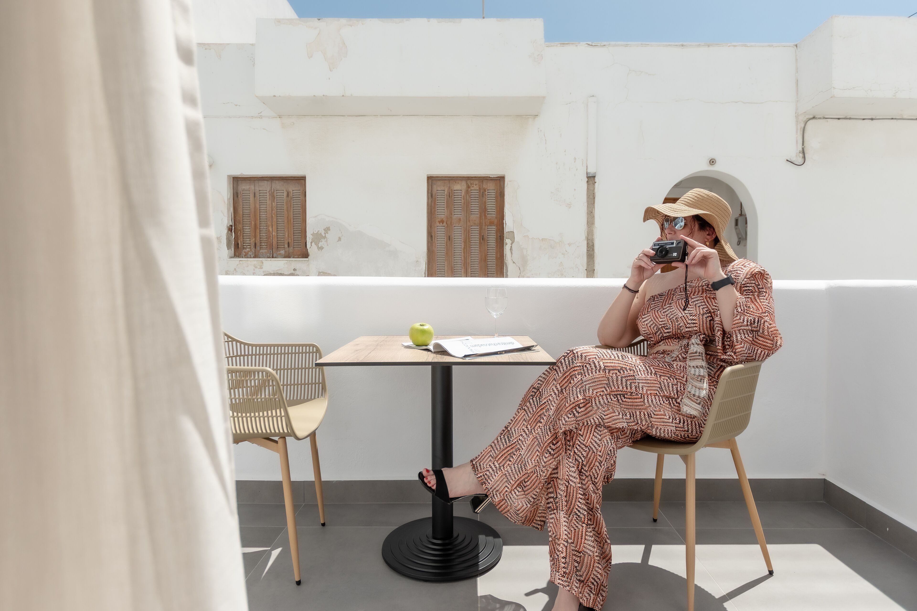 A person sitting on a chair at a table with a book and a glass of water, on a balcony with a white wall and wooden shutters.