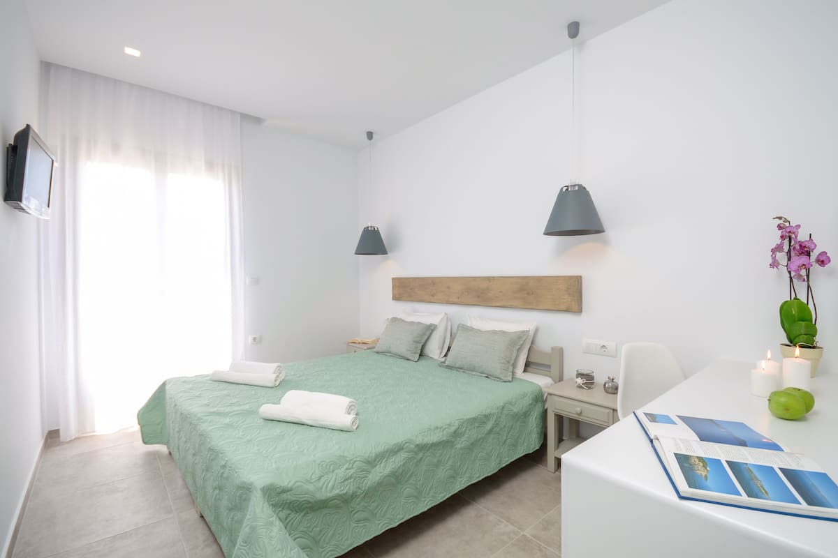 Hotellihuone, jossa on sänky, yöpöytä, lamppu ja pieni työpöytä, jonka päällä on lehti.