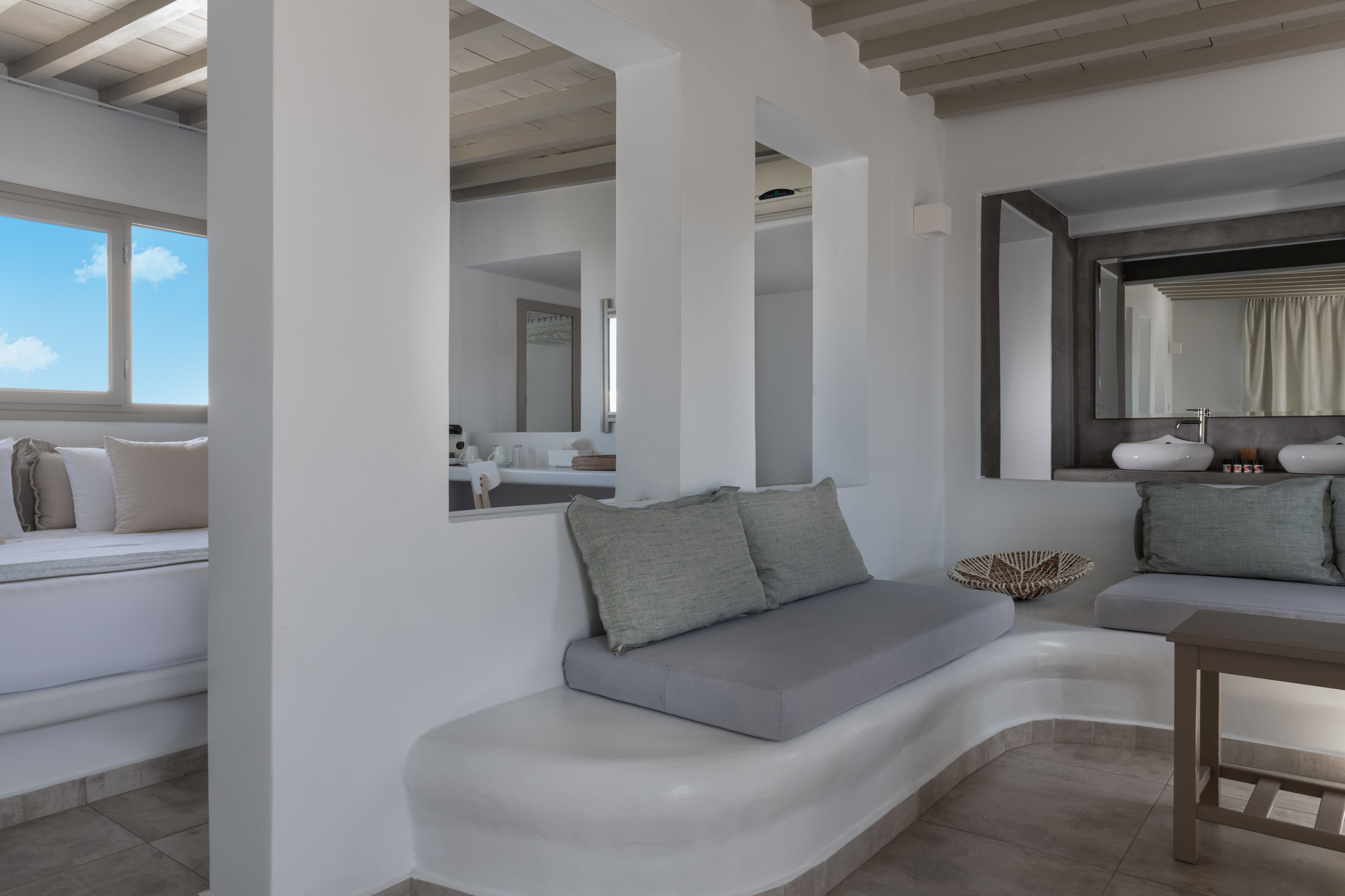 A modern living room with a curved white sofa, a small wooden table, and a large mirror reflecting a bathroom with a bathtub and white fixtures.