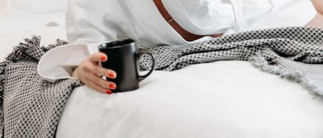 A person in a white bathrobe sitting on a bed with a black mug, a white pillow, and a gray blanket.