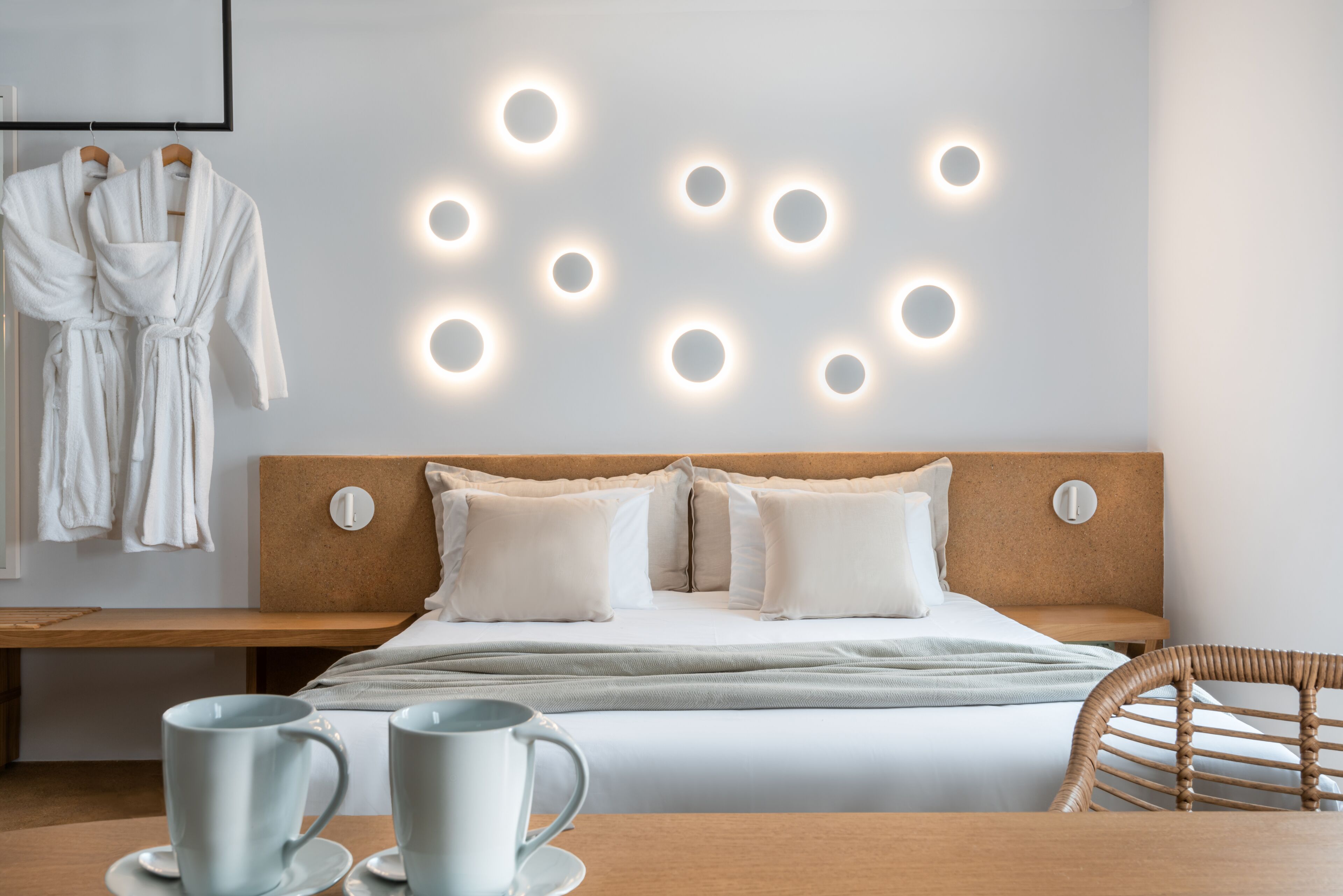 A hotel room with a bed, bedside table, two mugs, and a wall with circular light fixtures.