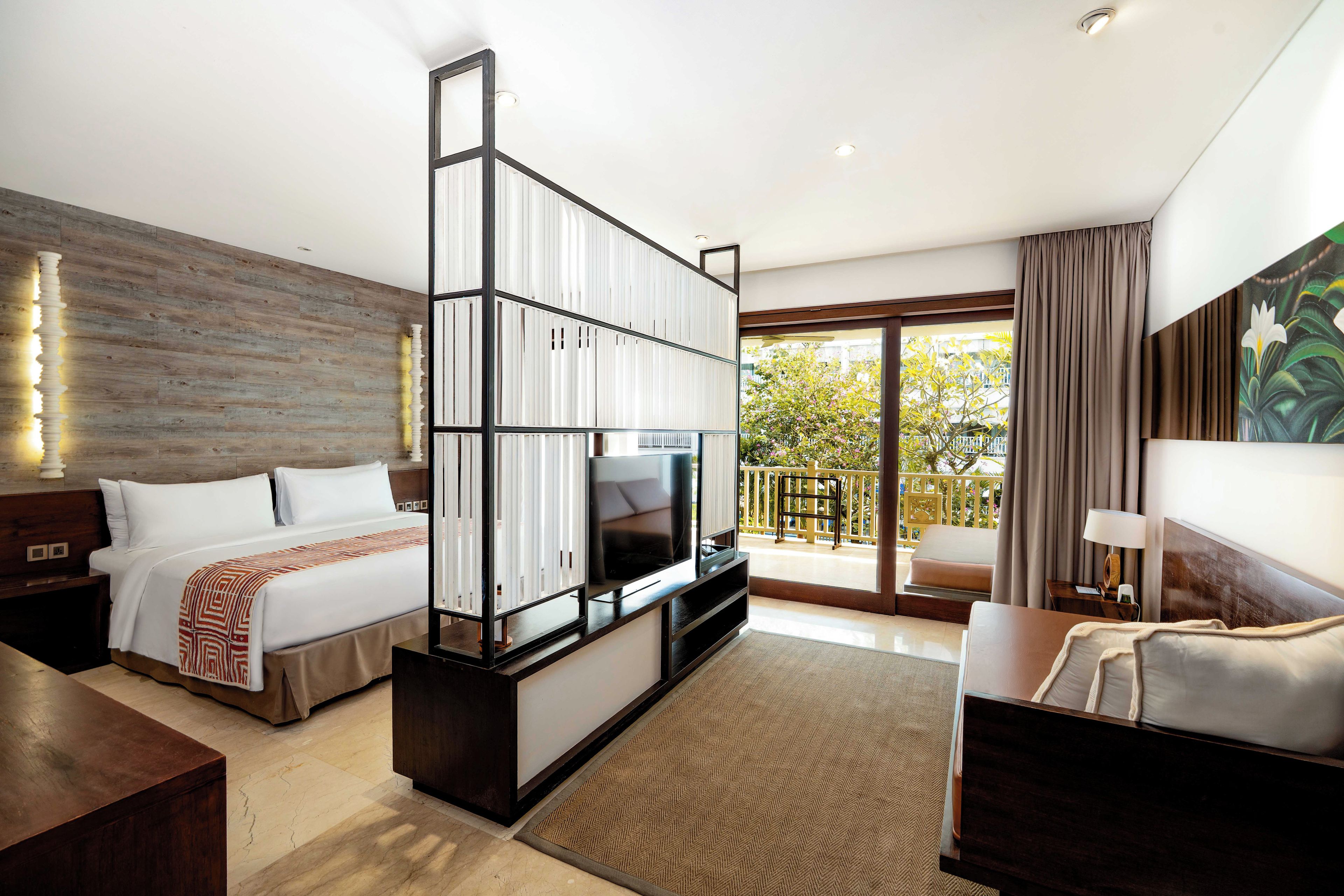 Ein Hotelzimmer mit Bett, Fernseher, Schreibtisch, Sofa und einem Balkon mit Blick auf Bäume und einen Zaun.