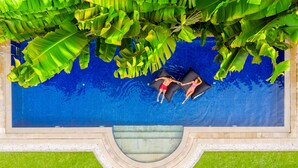 Vista aérea de uma piscina com duas pessoas deitadas na borda, rodeadas por folhagem verde exuberante.