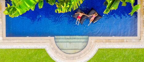 An aerial view of a swimming pool with two people lying on the edge, surrounded by lush green foliage.