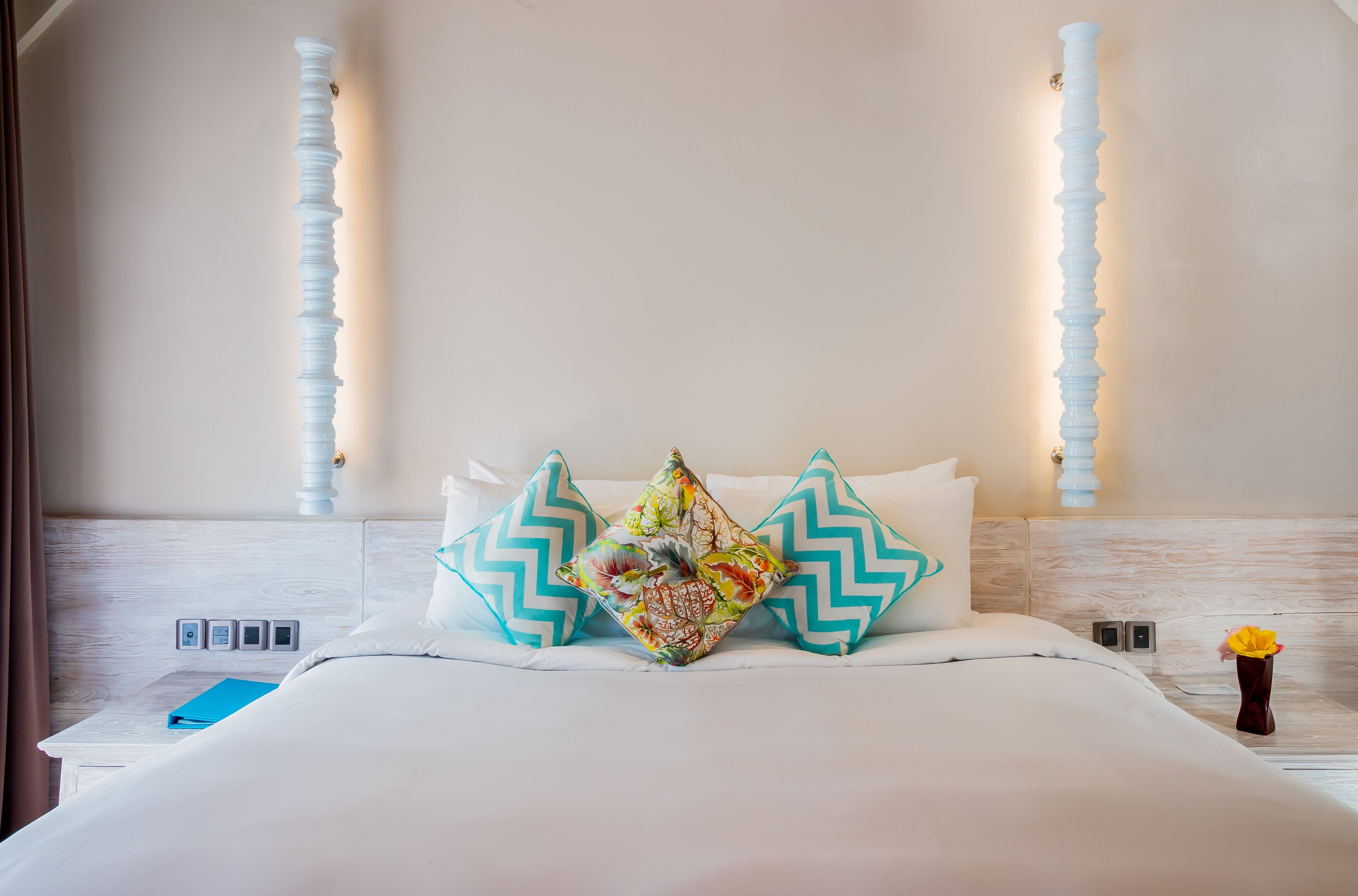 A neatly made bed with a floral pillow, a blue book, and a vase with yellow flowers.