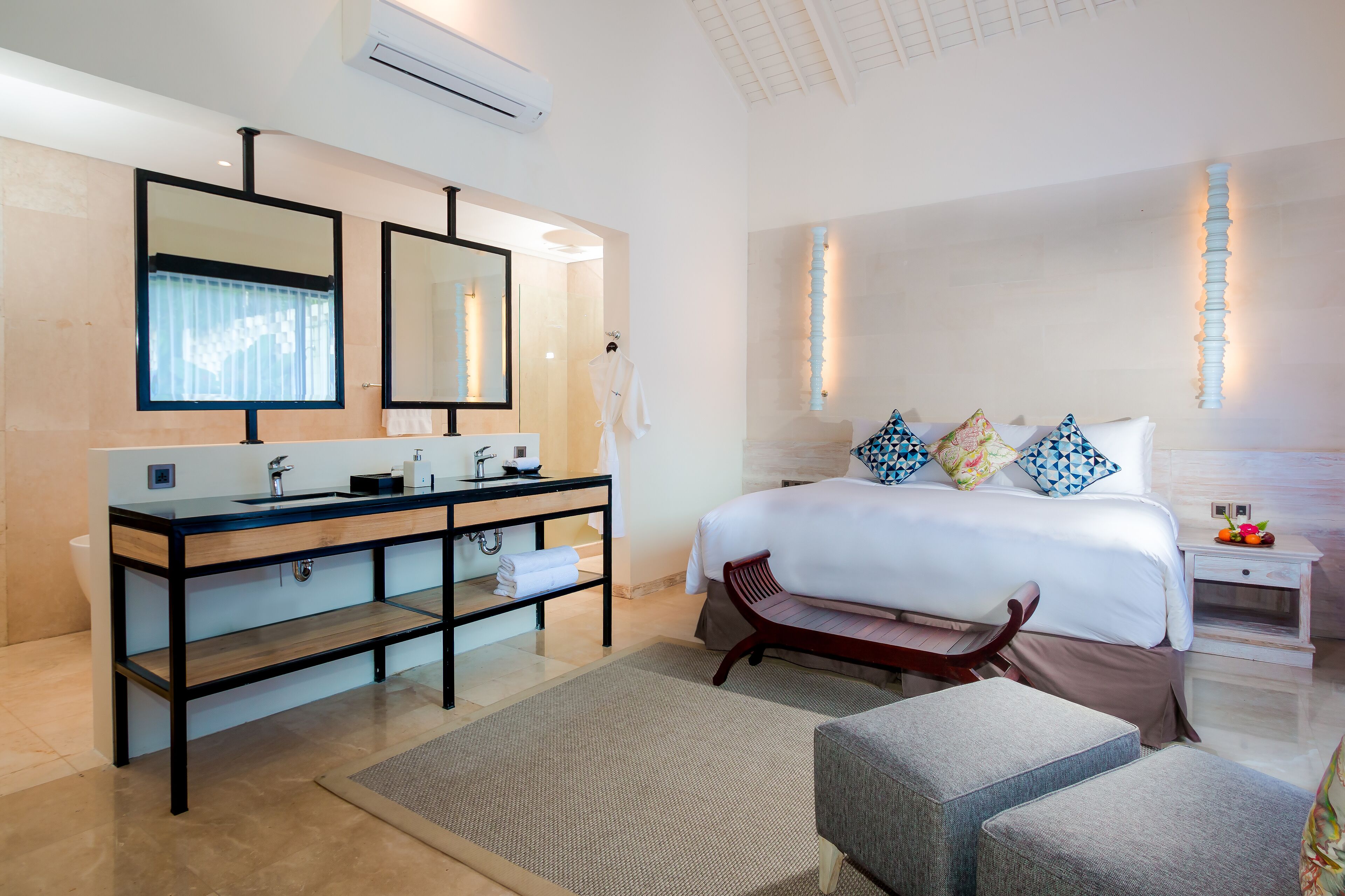 A modern hotel room with a large bed, a double sink, a mirror, and a bedside table with a fruit bowl.