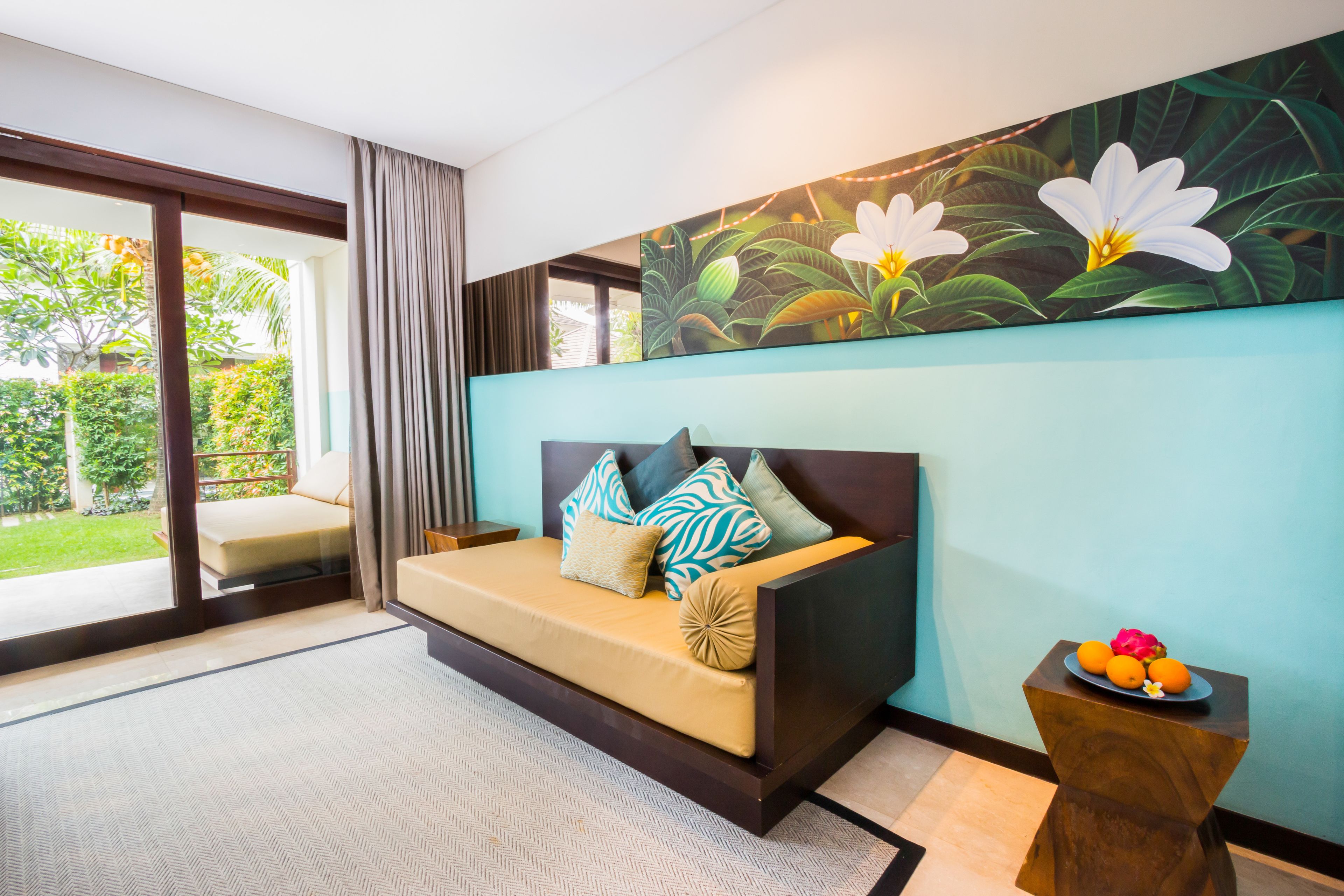 A modern bedroom with a sofa bed, a side table with fruit, and a large floral painting on the wall.