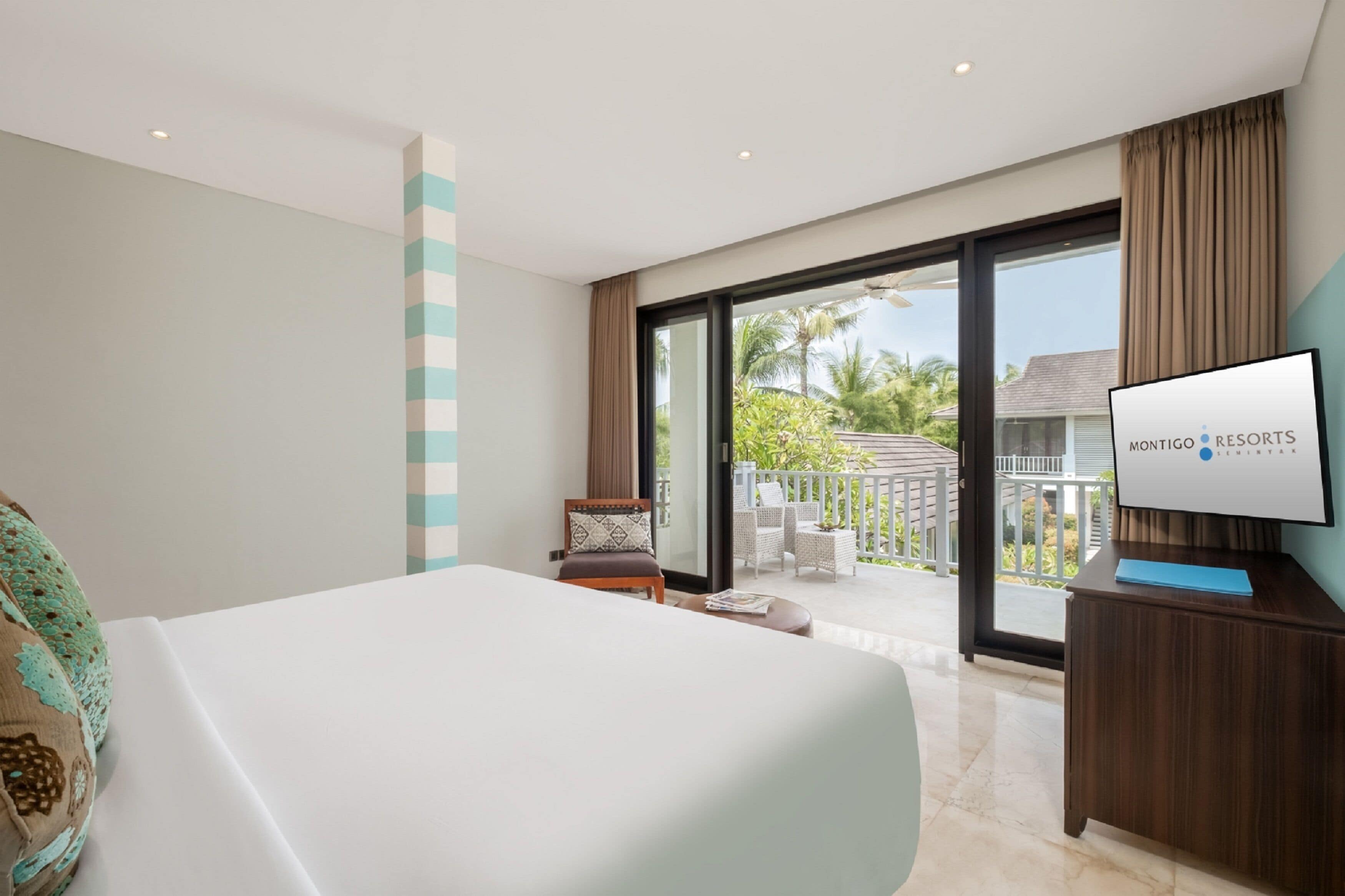 A hotel room with a large bed, a television on a wooden stand, a balcony with outdoor furniture, and a view of palm trees.