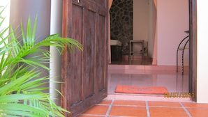 Uma porta de madeira com uma tranca de metal, um vaso de plantas e um chão de azulejos que levam a um quarto com uma parede de pedra e uma janela com padrão treliçado.