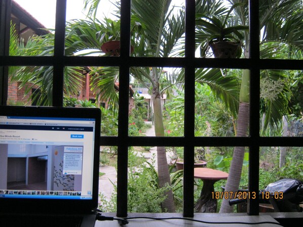 Um laptop sobre uma mesa, vista pela janela de um jardim com palmeiras e um edifício de tijolos.