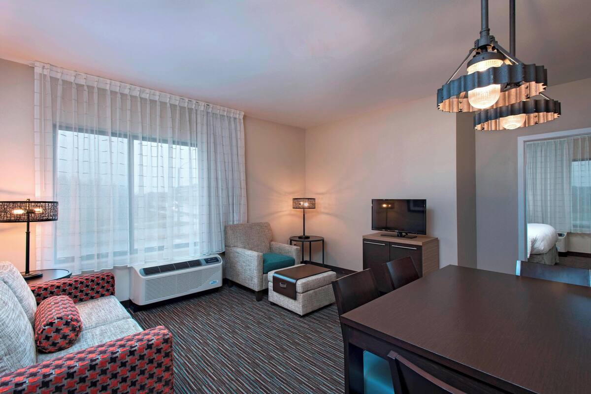 A hotel room with a sofa, armchair, ottoman, TV, dining table, and a unique chandelier.
