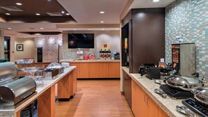 A hotel kitchen with buffet stations, a coffee machine, and a large flat-screen TV on the wall. - TownePlace Suites by Marriott Des Moines West/Jordan Creek (West Des Moines)