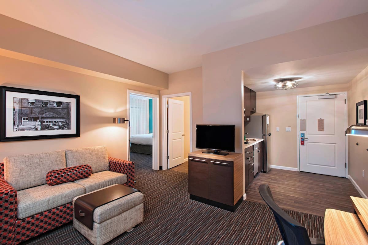A hotel room with a sofa, a television, a small kitchenette, and a framed picture on the wall.