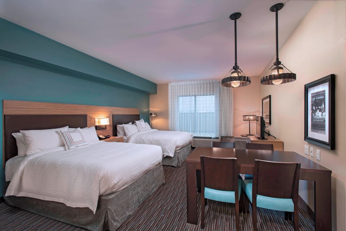 A hotel room with two beds, a dining table, chairs, a desk with a computer, and a framed picture on the wall.