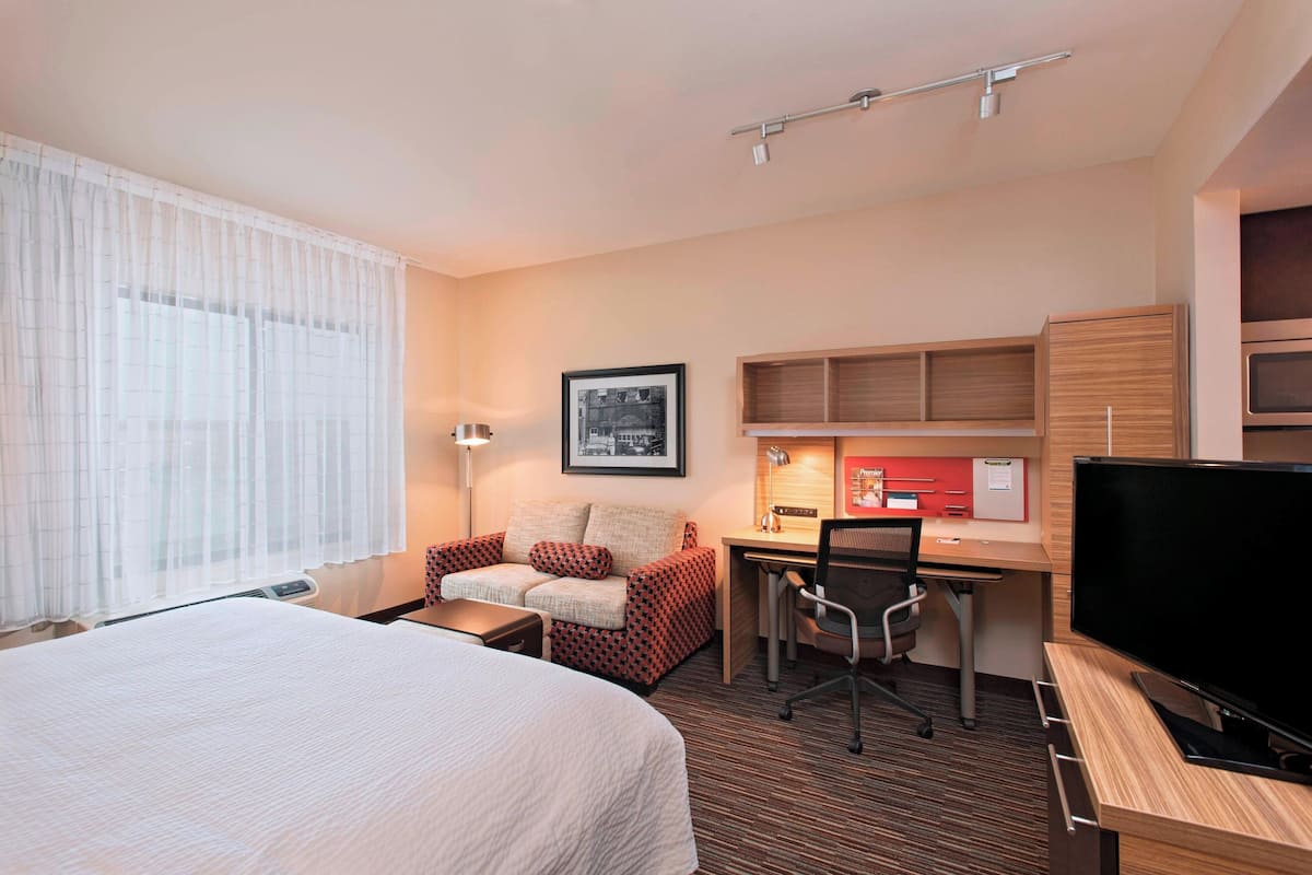 A hotel room with a bed, a desk with a computer, a chair, a TV, and a window with curtains.