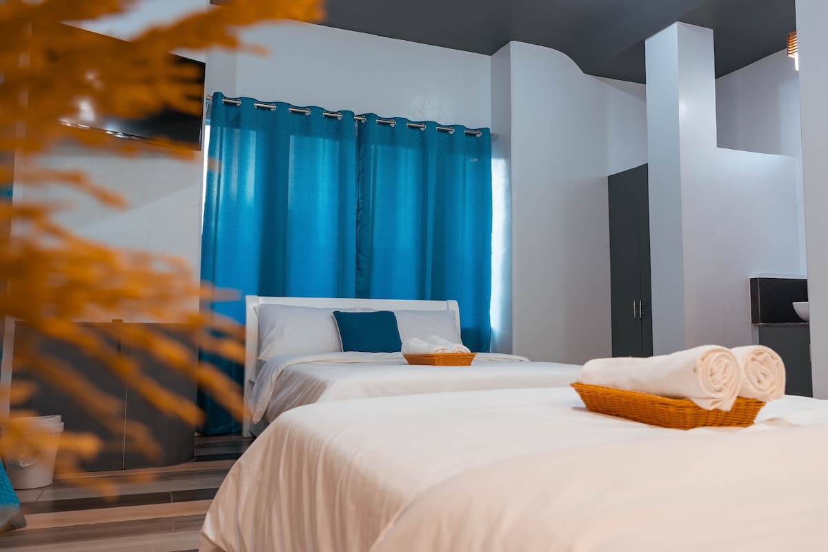 Habitación de hotel con dos camas, una cortina azul, ropa de cama blanca y una cesta con toallas.