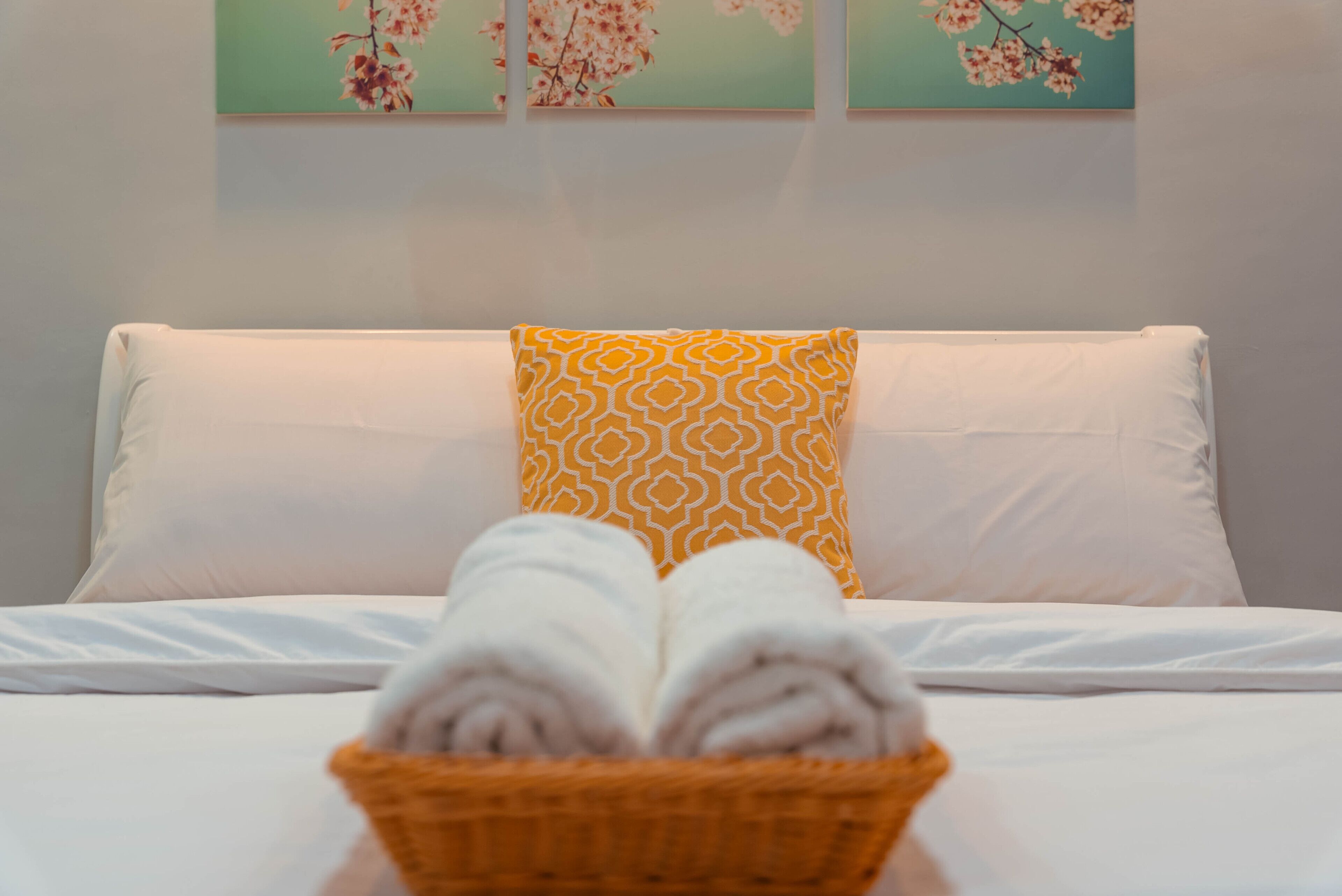 A bed with white linens, a yellow patterned pillow, and a basket of folded towels.