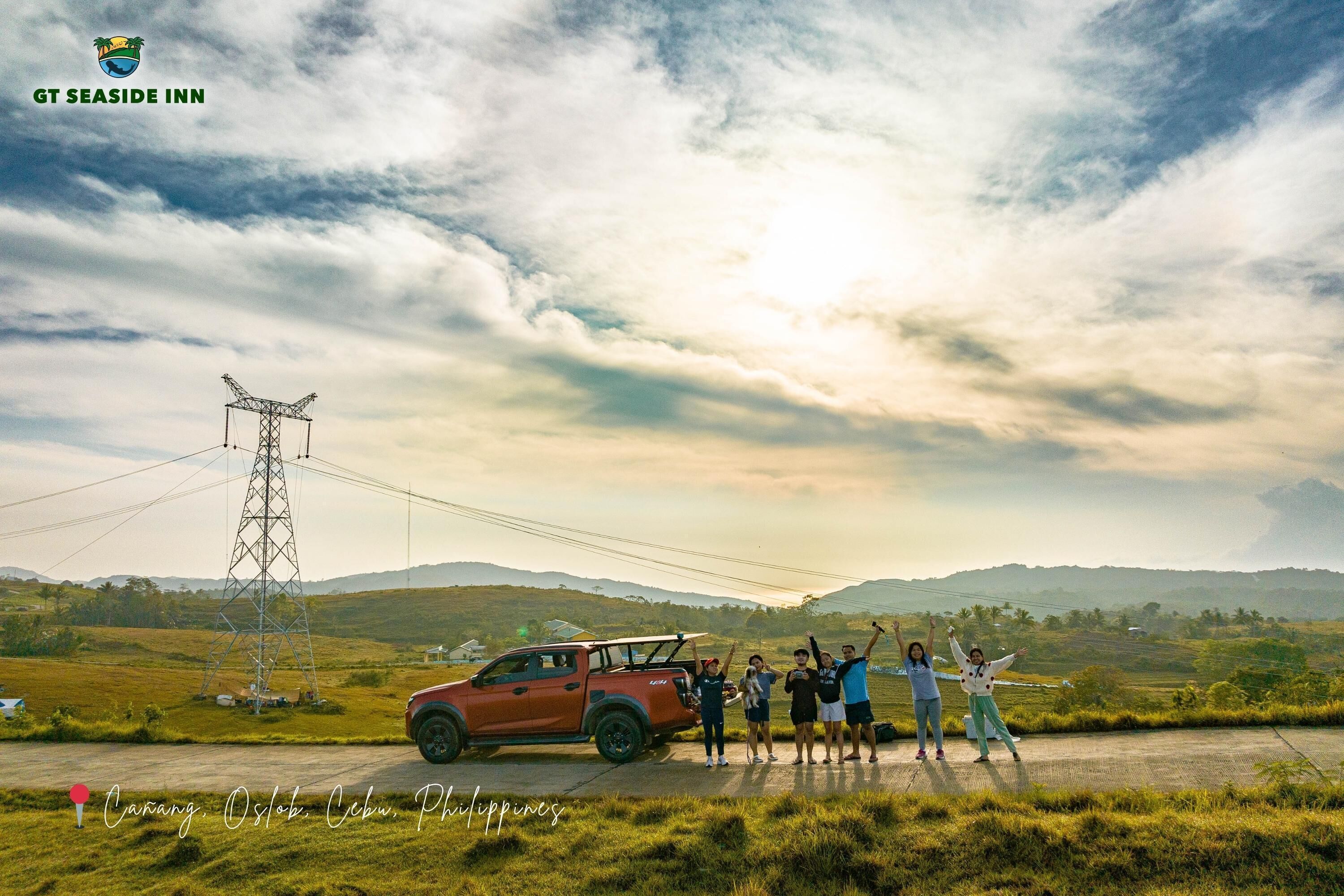 A group of people standing in front of a red vehicle on a rural road with a power line tower in the background.