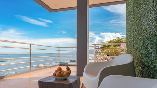 Un balcon avec vue sur la mer, un saladier rempli de fruits et des chaises blanches.