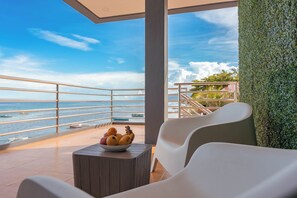 A balcony with a view of the sea, a bowl of fruit, and white chairs.