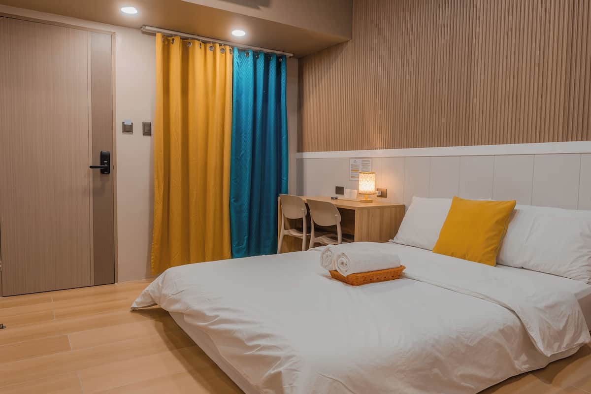 Una cama bien hecha con una toalla enrollada, una mesita con una lámpara y una silla en una habitación moderna con suelo de madera y cabecero de madera.