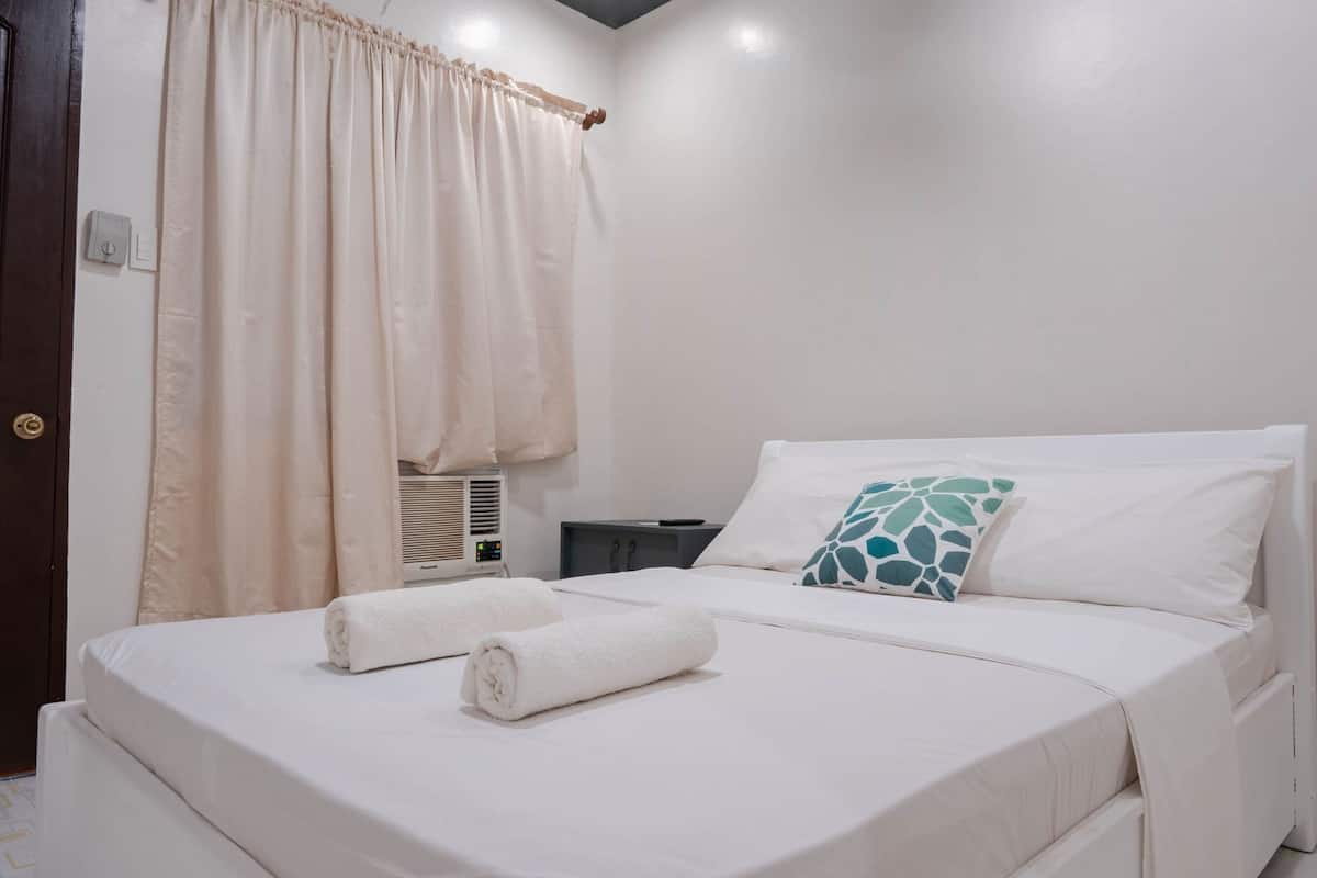 Un dormitorio con cama, mesita de noche, lámpara, ventana con cortinas y aire acondicionado.