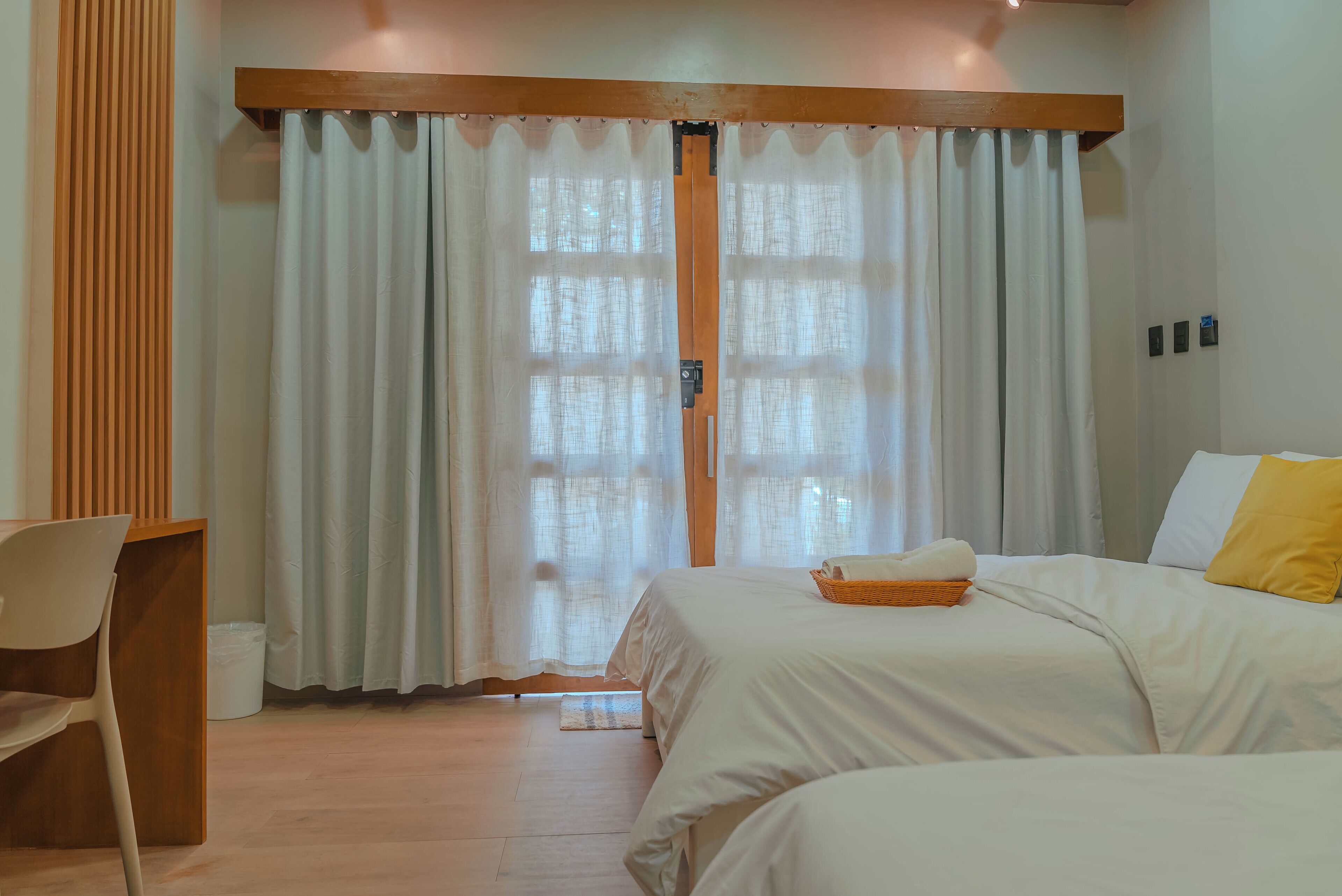 Une chambre avec deux lits, une tête de lit en bois, une chaise blanche et une fenêtre ornée de rideaux légers.