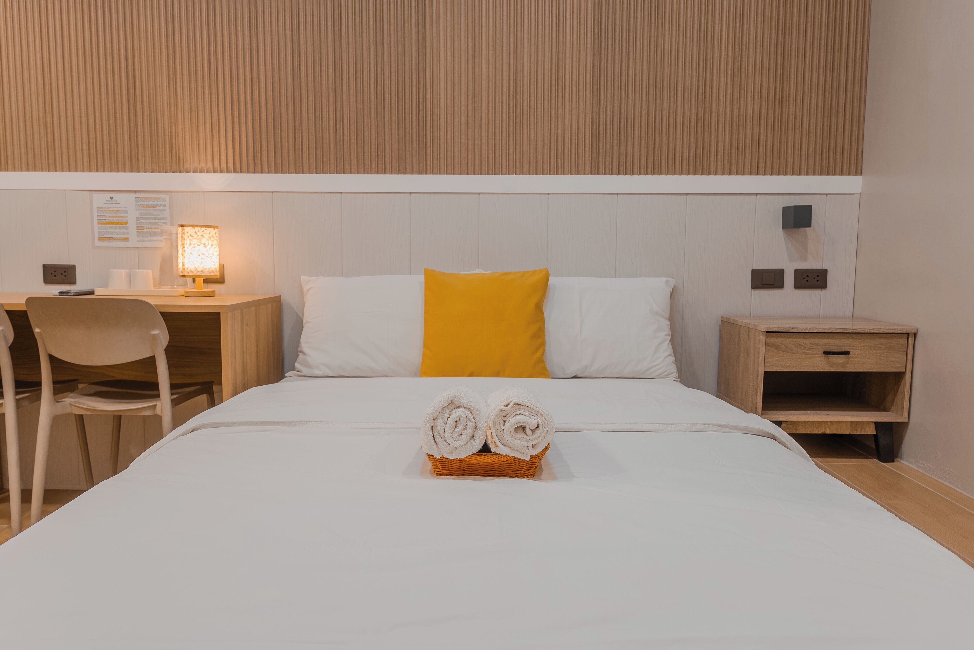 A neatly made bed with two rolled towels in a basket, a desk with a lamp, a chair, and a nightstand with a drawer.