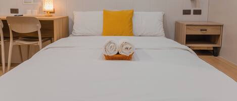 A neatly made bed with two rolled towels in a basket, a desk with a lamp, a chair, and a nightstand with a drawer.