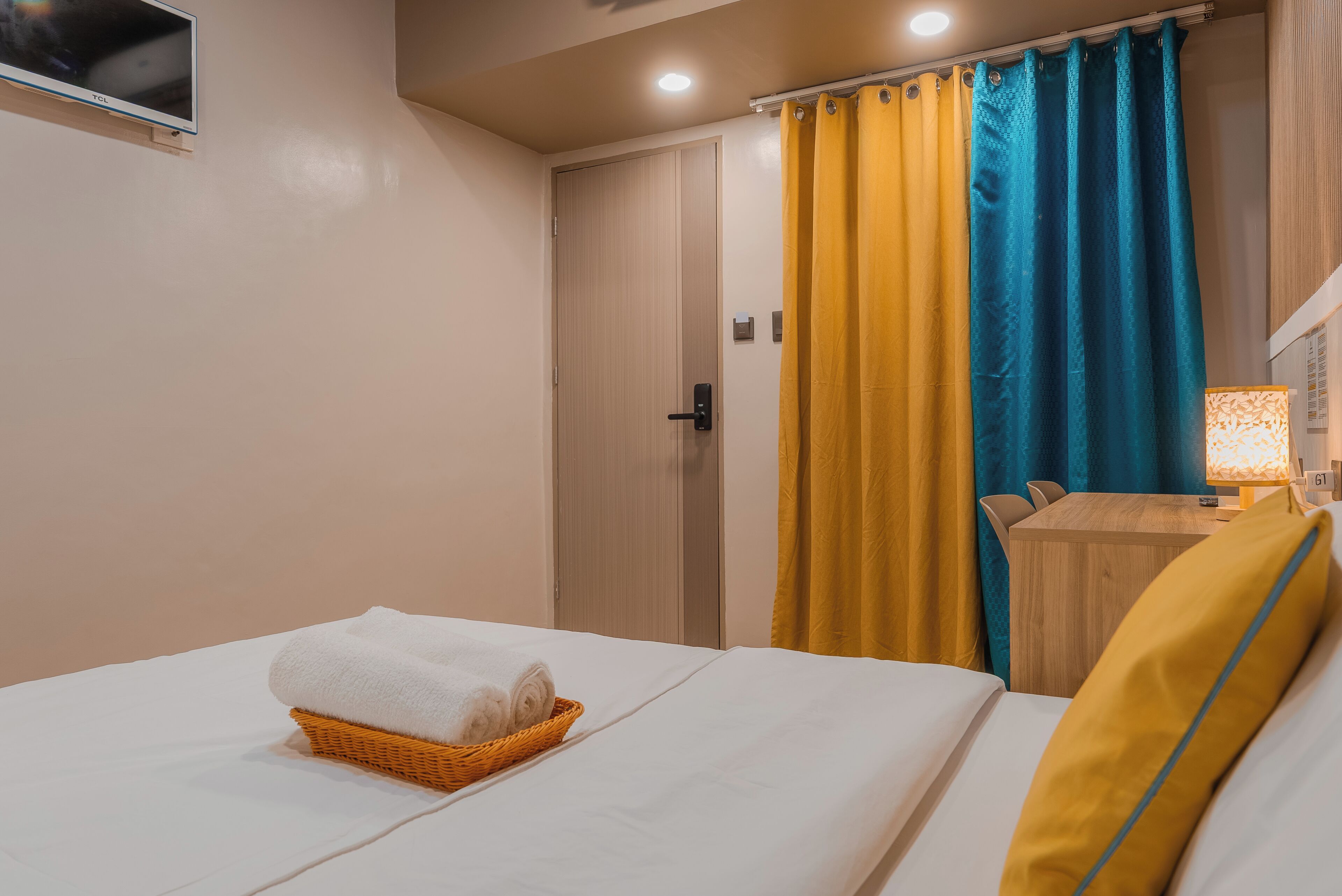 Habitación de hotel con una cama, una puerta de madera, una cortina amarilla y azul, una mesita con una lámpara y un televisor montado en la pared.