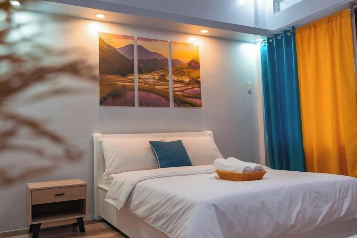 A neatly made bed with a white comforter and a basket of towels, a bedside table, and a wall-mounted painting of a landscape.