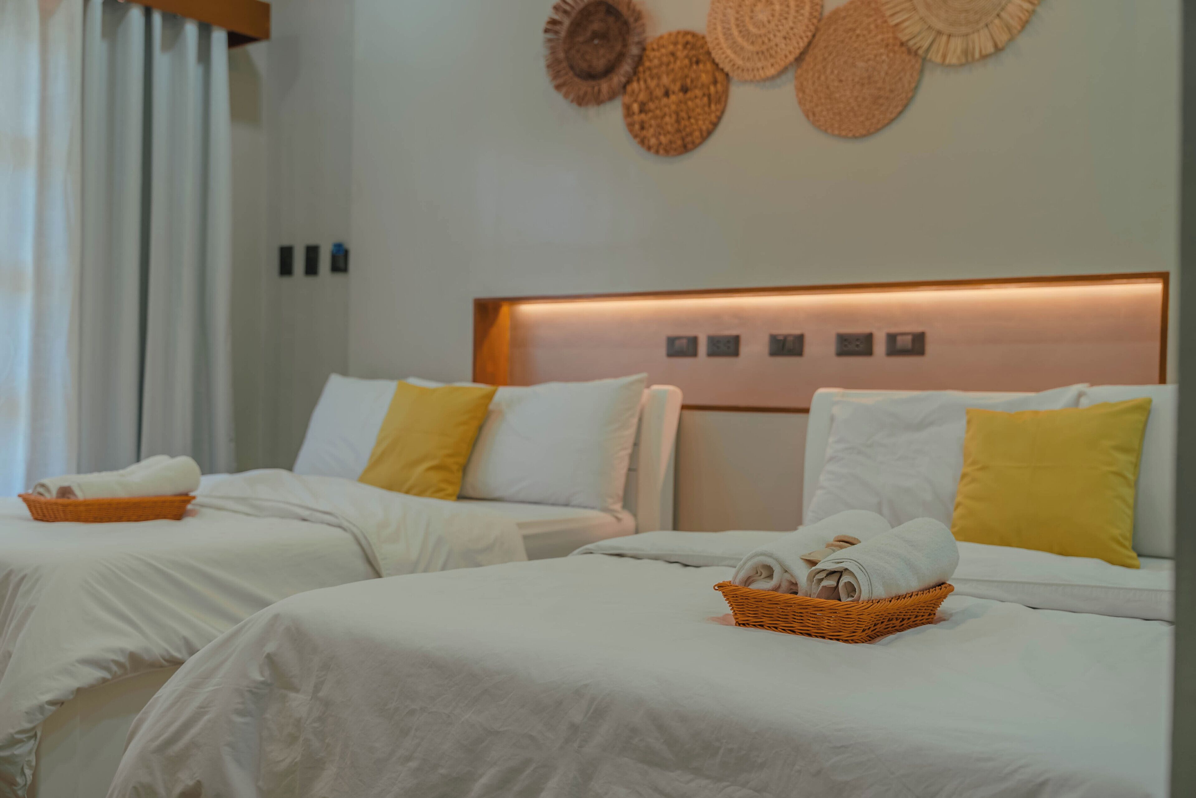 A hotel room with two beds, white bedding, yellow pillows, and a woven wall decoration.