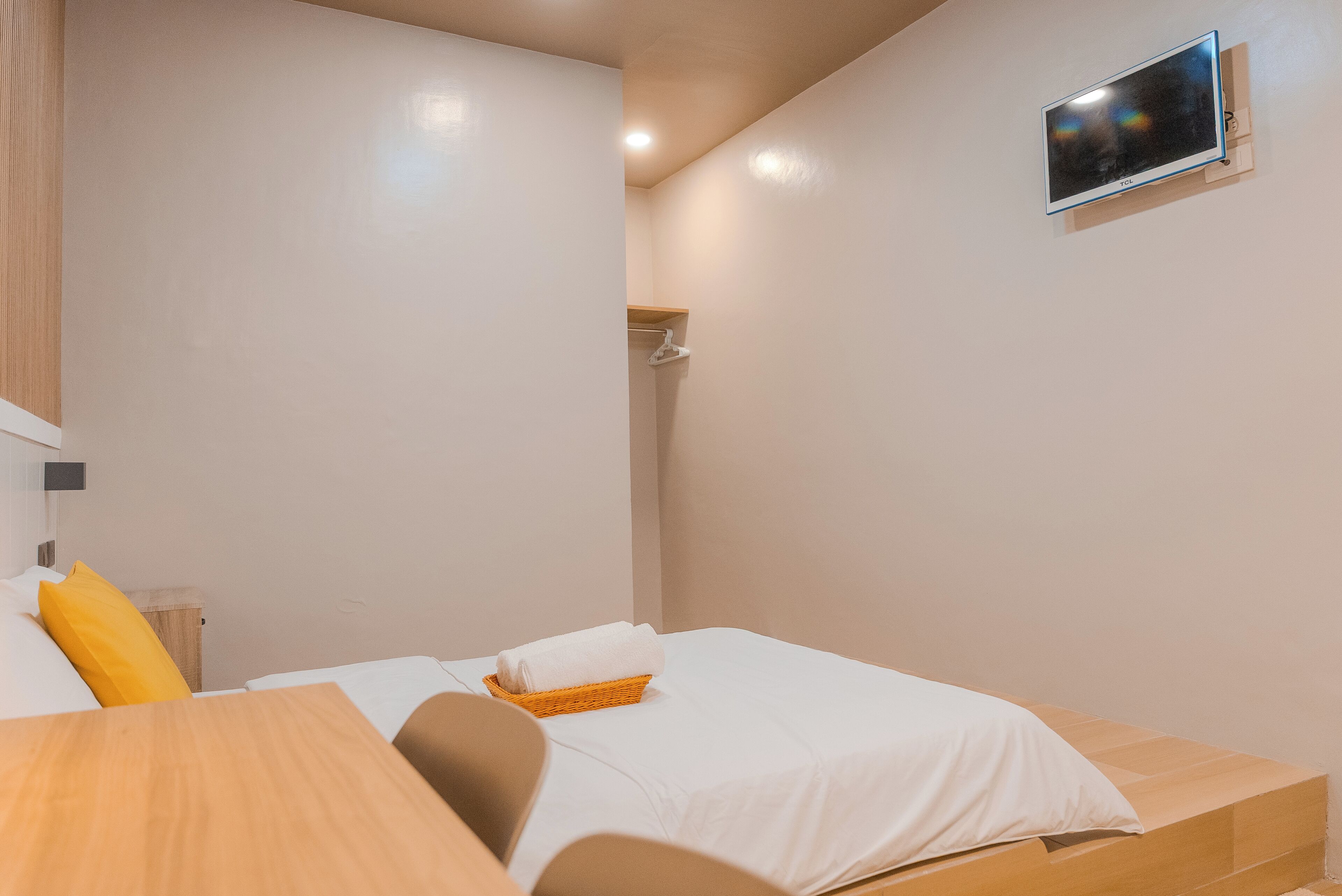 A bedroom with a bed, a wooden nightstand, a TV mounted on the wall, and a small shelf.