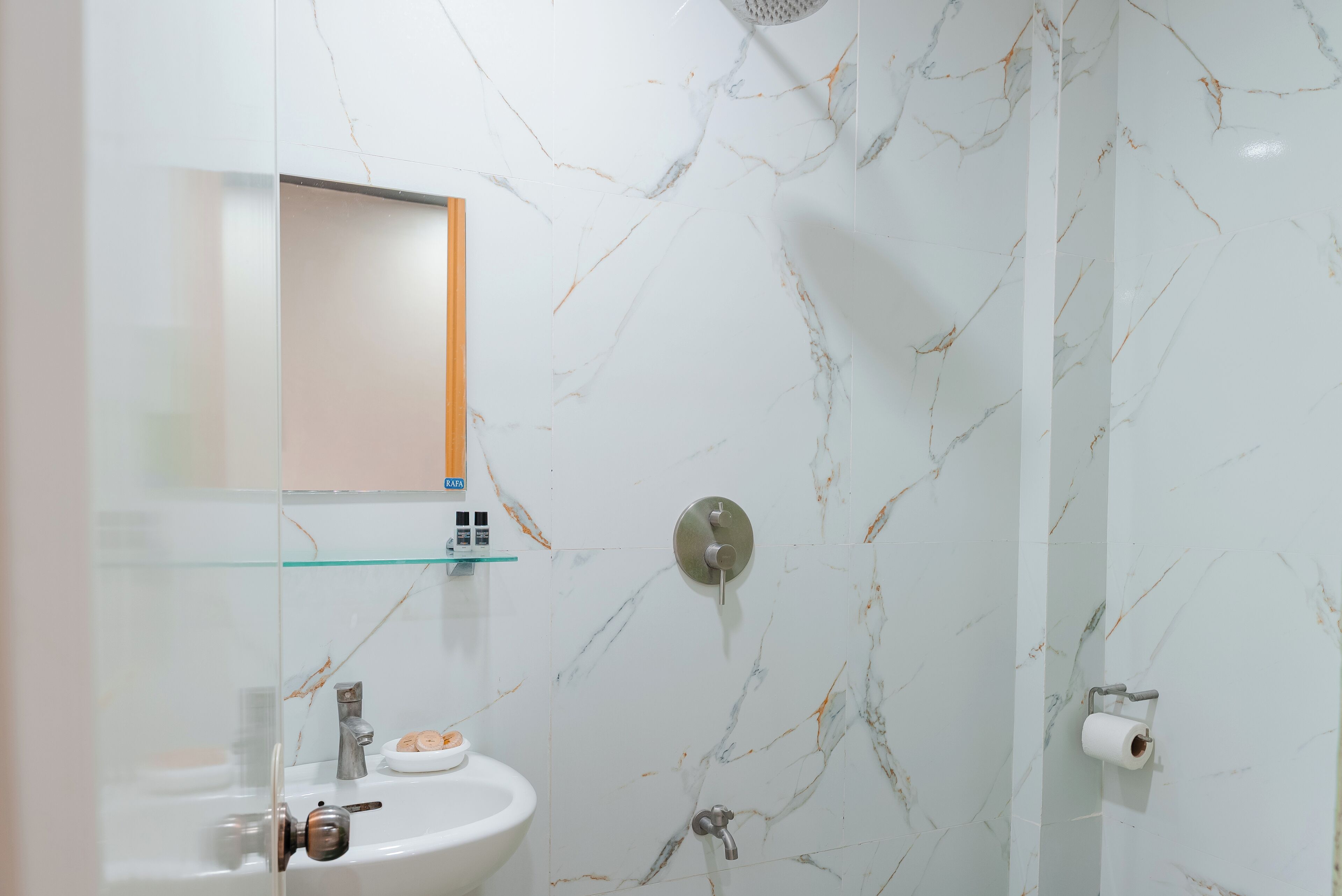 A bathroom with a marble wall, a glass shelf with toiletries, a sink, and a shower.