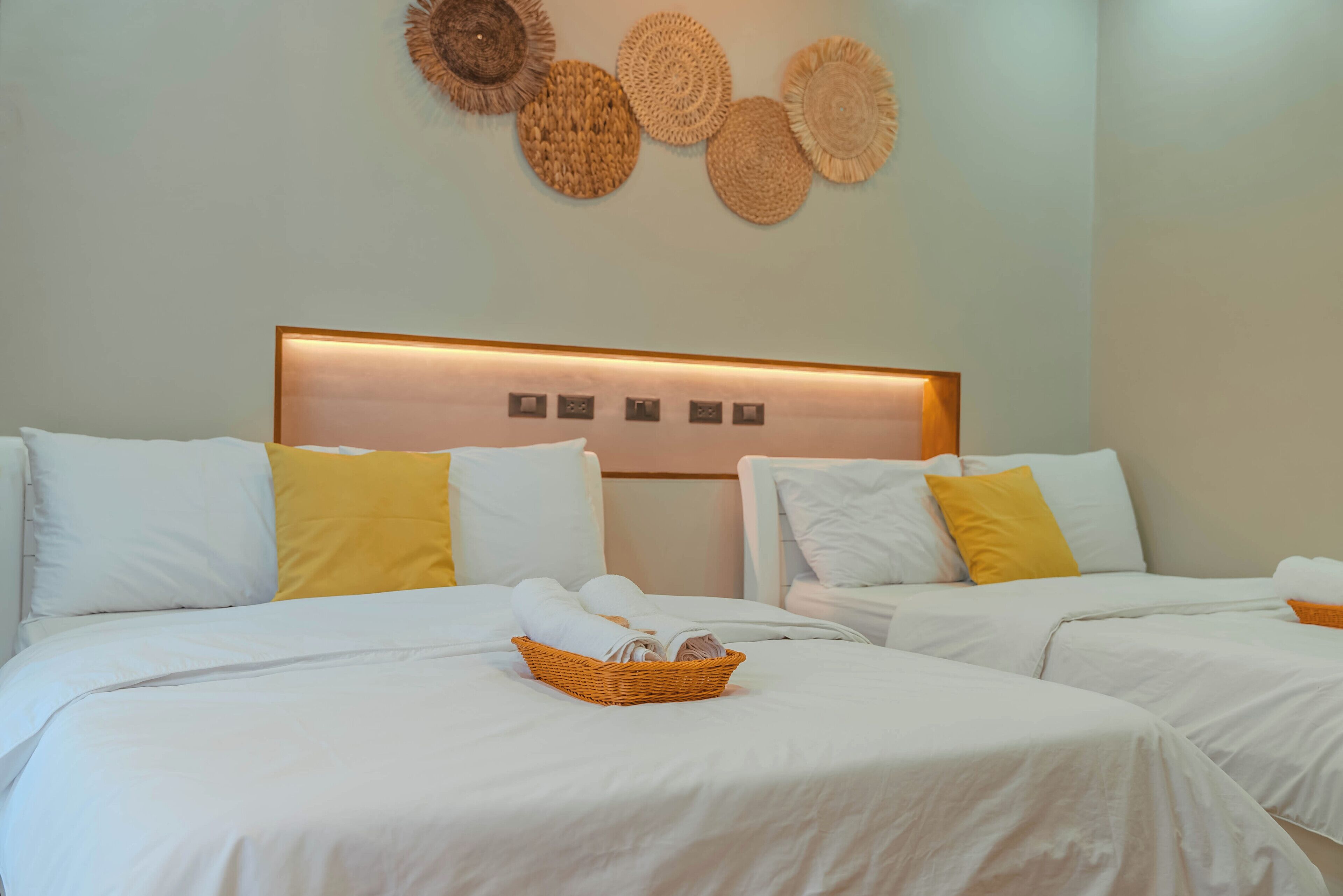 Habitación de hotel con dos camas, ropa de cama blanca, almohadas amarillas, cabecera con panel de luz y una obra de arte tejida colgada en la pared.