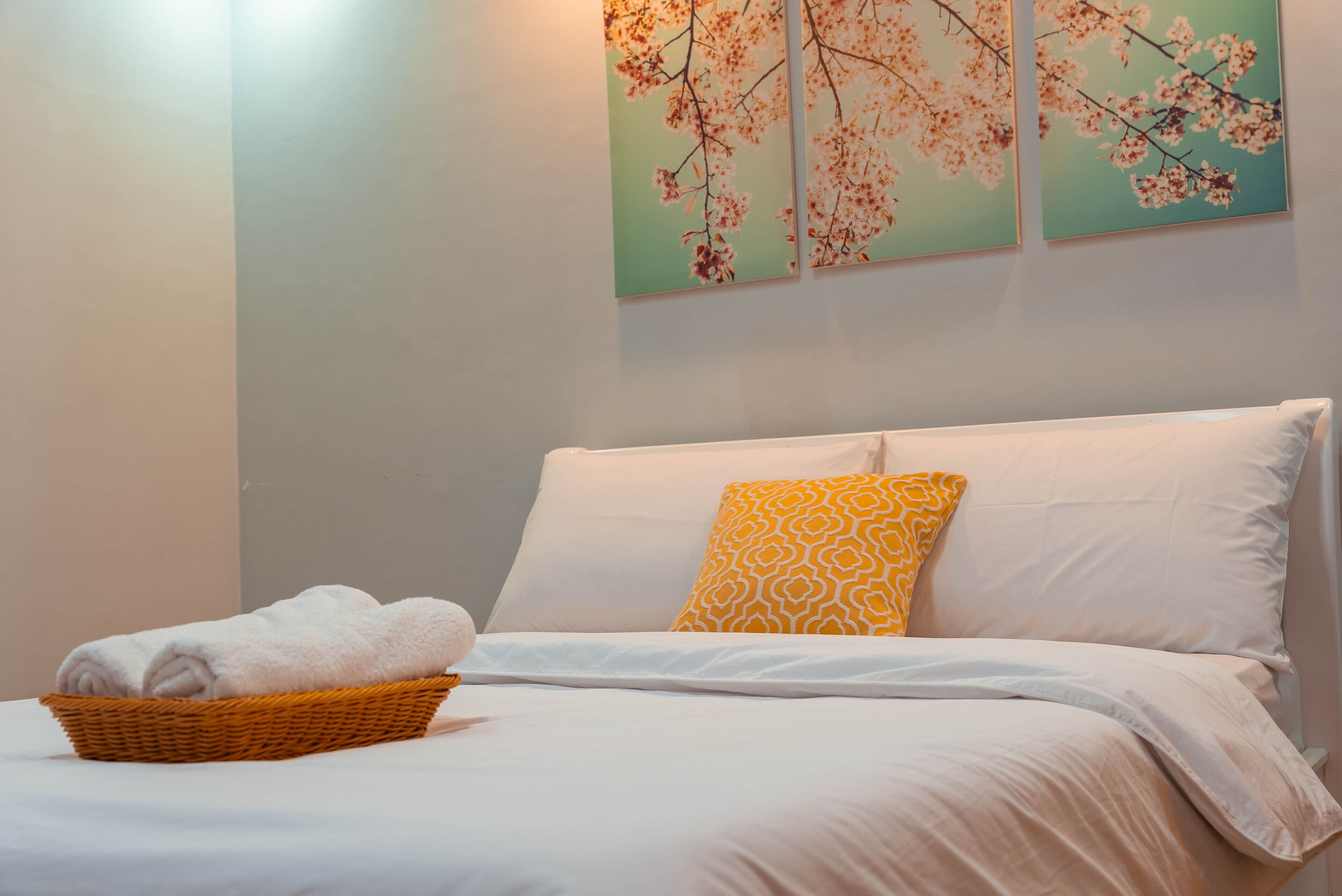 Un lit bien fait, agrémenté d’oreillers blancs et à motifs, un panier rempli de serviettes pliées et deux tableaux floraux accrochés au mur.