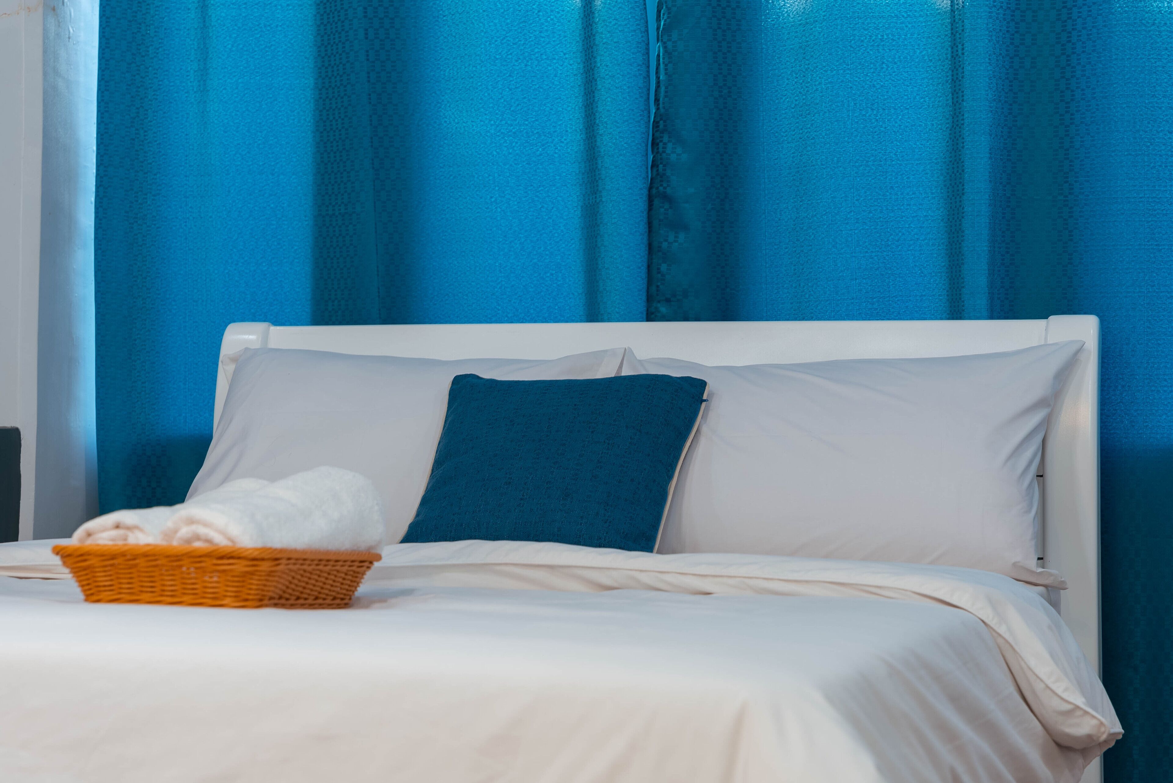 Una cama con ropa blanca, una almohada azul y una cesta de toallas.