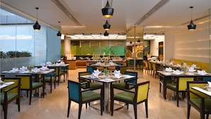 Un espace repas moderne avec plusieurs tables et chaises, une cuisine à l’arrière-plan et de grandes fenêtres.