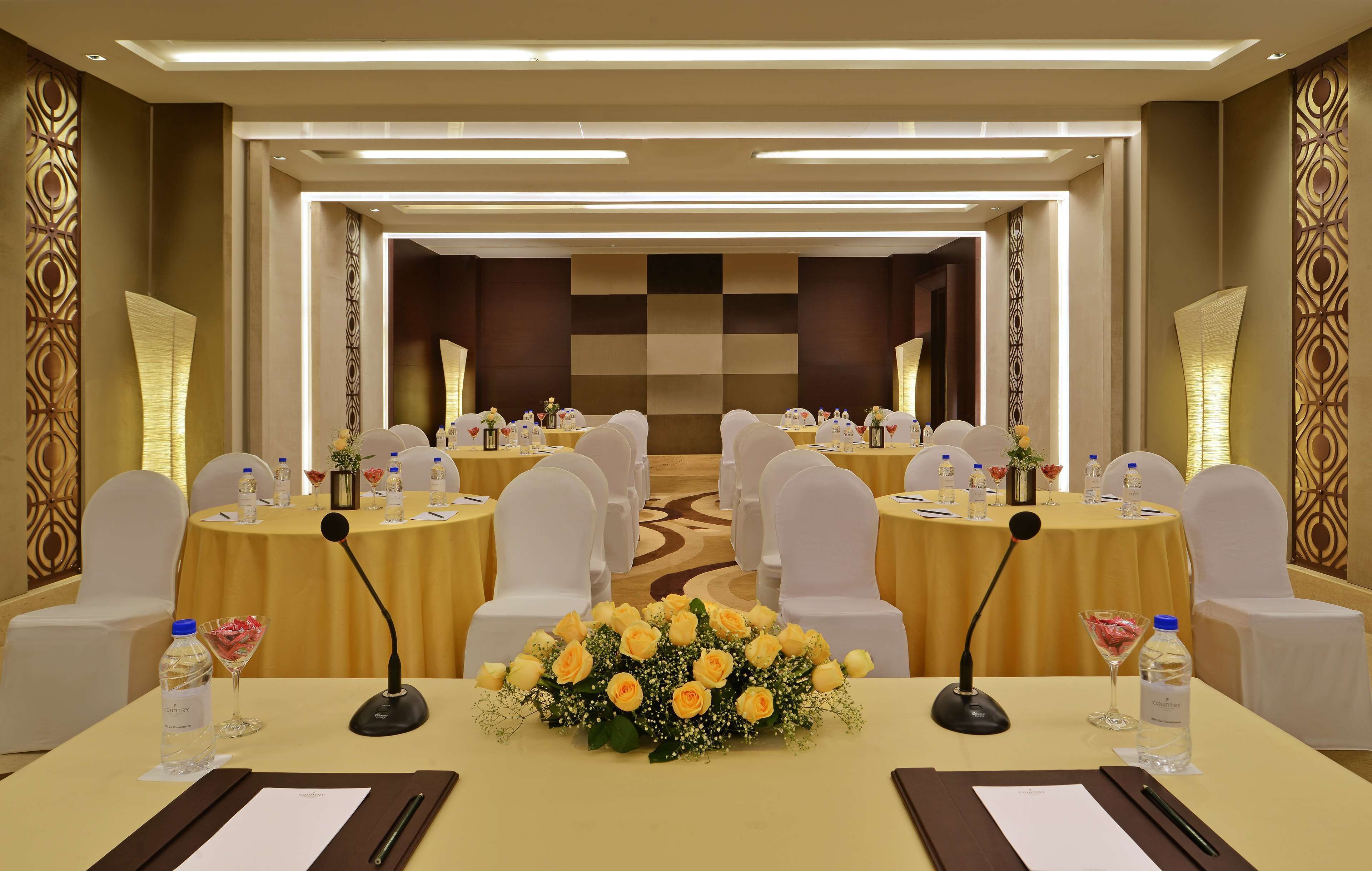 A conference room with round tables, white chairs, microphones, water bottles, and floral centerpieces.
